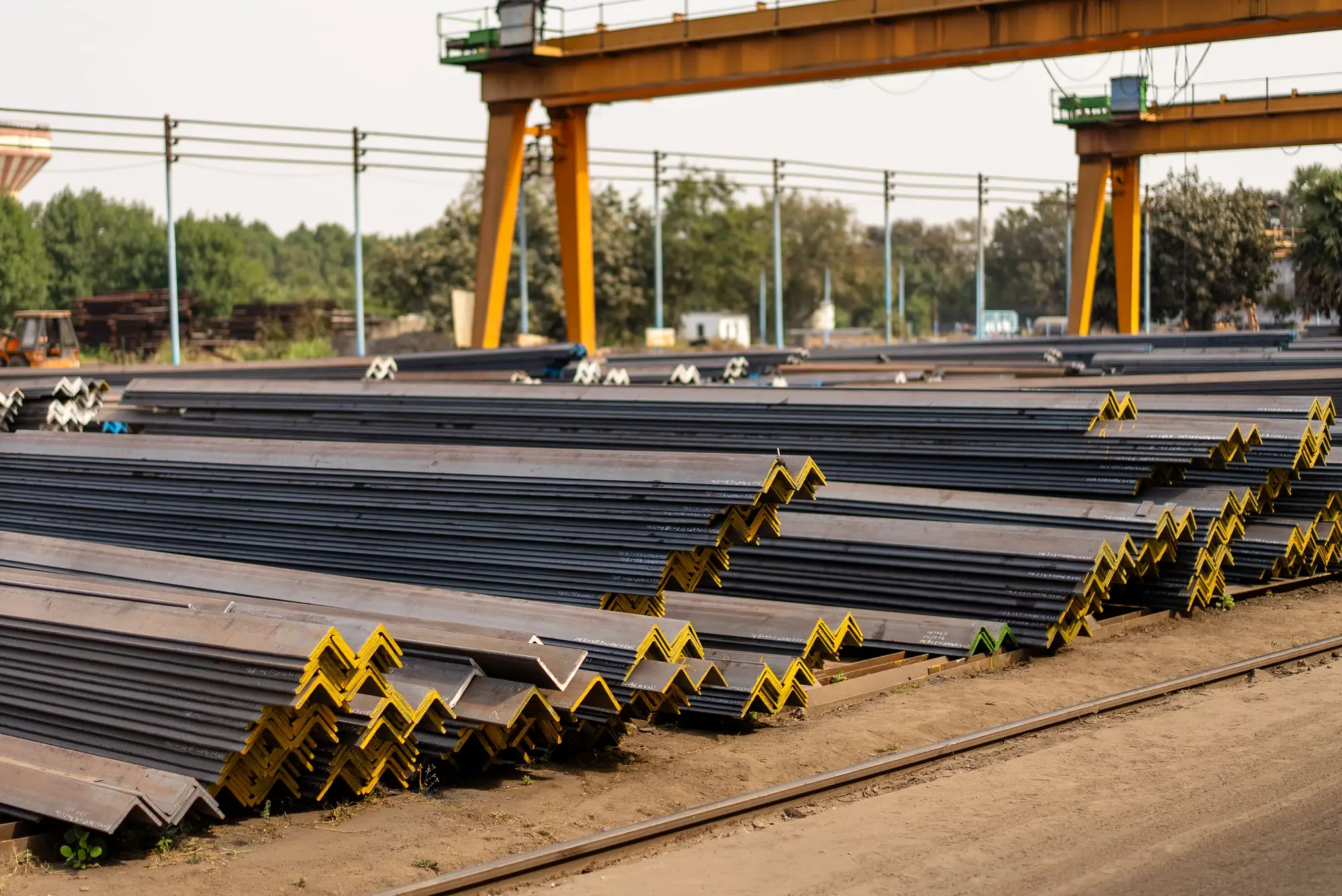 Steel angle stock stacked in rows across an open yard under a gantry crane